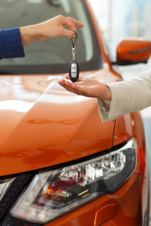 A key fob is handed off from a car rental concierge to a driver in front of an orange car.